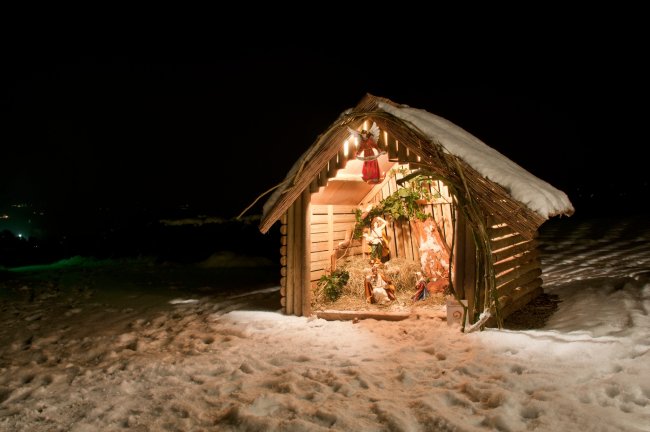 Een sfeervolle kerststal in een besneeuwde houten hut, verlicht tegen een donkere nachtelijke achtergrond, met traditionele kerstfiguren binnenin.