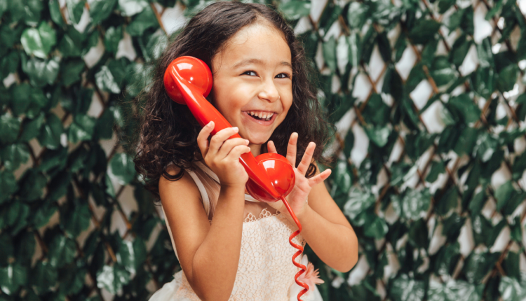 Een lachend meisje met een rode speelgoedtelefoon, tegen een achtergrond van groene bladeren.