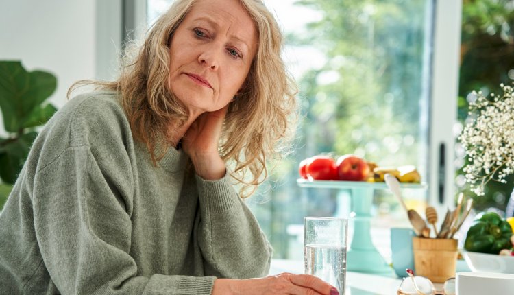 Vrouw kijkt zorgelijk en zit met de hand in de nek en een glas water aan tafel