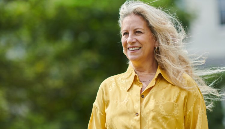 Vrouw met geel shirt en mooi blond haar kijkt lachend uit over haar tuin