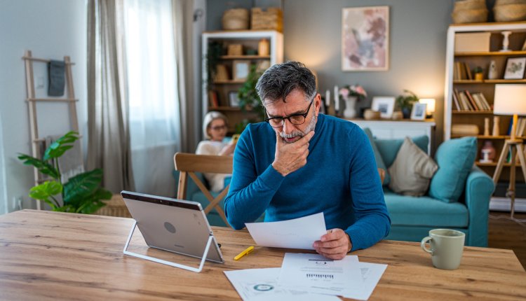 Man is thuis bezig met financien, vrouw op de achtergrond op de bank aan het lezen