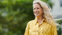 Vrouw met geel shirt en mooi blond haar kijkt lachend uit over haar tuin