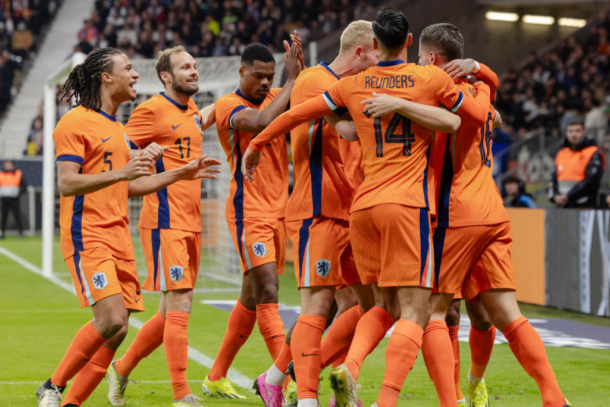 Nederlandse voetballers vieren een doelpunt tijdens een wedstrijd. Ze dragen oranje shirts met blauwe accenten. Een speler, te herkennen aan rugnummer 14, omhelst zijn teamgenoten terwijl anderen juichen. Op de achtergrond zijn toeschouwers en stadionpersoneel te zien.