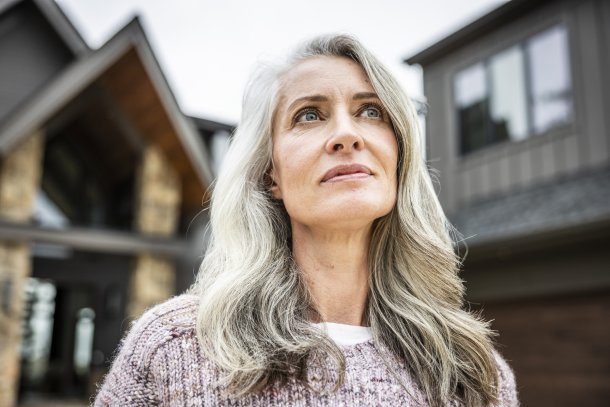 Knappe oudere vrouw met prachtig golvend grijs haar staat voor haar huis en kijkt bedachtzaam